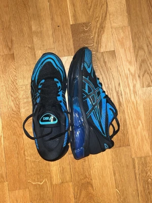 Asics gel 360 - Säljer ett par snygga ASICS sneakers i svart och blått med coola detaljer och mönster på sidan. Skorna har snörning och en platt sula med extra dämpning, perfekt för dig som gillar sportig stil. Materialet är syntet och mesh för bra andning.