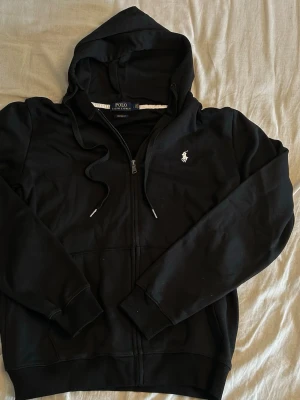 Svart Ralp Lauren zip  - En svart Ralph Lauren Zip med vit logga. Köpt från Facebook marketplace som ny och bara test använd. Köpte den men passade inte jätte bra. Storlek L men passar nog M för jag är 187 och va något för stor för mig så lägger ut den som M så mellan 178-185 borde den passa bra. Finns ångerrätt så går att köpa och skicka tillbaka om den inte passar eller något annat beskymmer så länge det köps via appen. Mvh