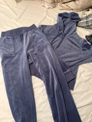 Blå mjukisbyxor från Juicy Couture - Säljer ett par blå mjukisbyxor från Juicy Couture med bred resår i midjan och snörning. Byxorna har fickor med lock och silverfärgade knappar samt broderad JC-logga på benet. Perfekt för chill dagar och snyggt matchande med hoodie. Byxorna är i xs och tröjan i S