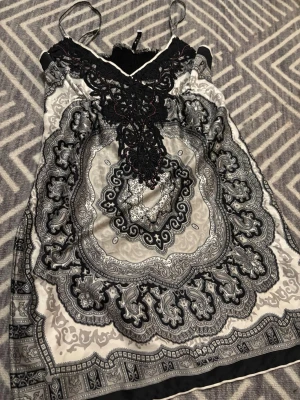 Svart och silver mönstrad klänning - Snygg klänning med tunna axelband och ett detaljerat svart spetsmönster vid halsen. Toppens tyg har ett paisleyinspirerat mönster i svart och silvertoner, och materialet är glansigt och känns lyxigt. 