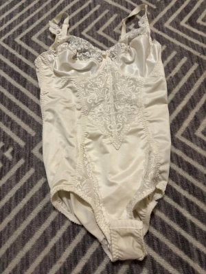 Vit spetsdetaljerad body i satin - Snygg vit body med tunna axelband och vackra spetsdetaljer framtill. Materialet är glansigt satin och spets och en liten rosett mellan kuporna. 