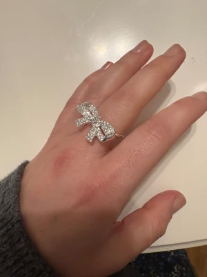 Glittrig rosett-ring i silver - Säljer en snygg ring i silverfärg med stor rosett framtill, täckt av glittrande stenar som ger extra bling. Ringen har en bred och stabil form, perfekt för dig som gillar statement-smycken och vill sticka ut. Rosetten är detaljrik och fångar ljuset superfint.