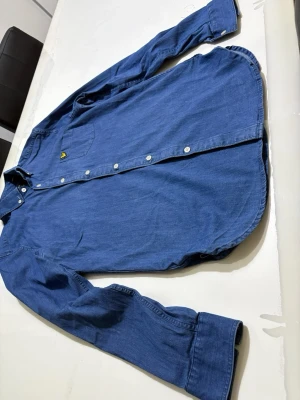 Blå skjorta från Lyle & Scott - Snygg blå skjorta från Lyle & Scott med klassisk button-down krage och vita knappar. Bröstficka med den ikoniska gula örnloggan ger en clean och stilren look. Skjortan är i mjuk bomull och har lång ärm, perfekt till jeans eller chinos. Storleken är S men passar även M.