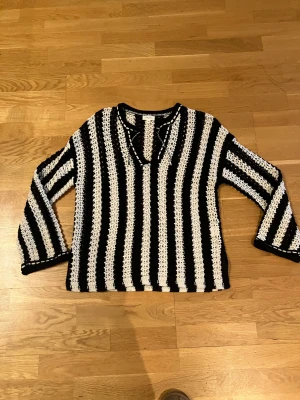 Svartvit randig stickad tröja - Säljer en svartvit stickad tröja med vertikala ränder och v-ringad hals med snörning. Tröjan har långärmade ärmar och ett luftigt, grovstickat material som ger en cool och avslappnad vibe. Perfekt för dig som gillar retro och statement-plagg.