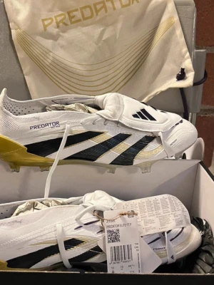 Adidas Predator Elite FT fotbollsskor - Adidas Predator Elite FT är grymma fotbollsskor i vitt med svarta och guldiga detaljer. Snygg design med snörning, låg profil och mönstrad sula för max grepp på planen. Skorna har en platt sula med dobbar och är tillverkade i syntetmaterial för lätt känsla och bra passform.