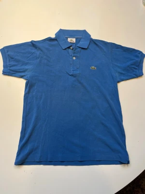 Blå piké från Lacoste med krage - Säljer en klassisk blå pikétröja från Lacoste med den ikoniska krokodilloggan broderad på bröstet. Har använts under sommaren men har inga hål eller synliga märken. Skriv vid frågor så svarar jag så snabbt jag kan😇. Pris kan diskuteras!