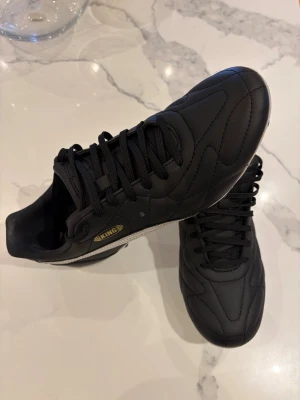 Puma King svartvita fotbollsskor - Säljer ett par klassiska Puma King fotbollsskor i svart skinn med vita detaljer och guldigt King-märke på sidan. Skorna har snörning, rund tå och dobbar för gräsplan. Perfekta för dig som vill ha stilrena och tidlösa fotbollsskor med grym känsla på planen.