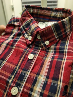 Rutig skjorta från Denim Day, stl 128 - Snygg rutig skjorta från Denim Day i rött, marinblått och beige. Klassisk button-down krage, vita knappar och svart dödskallebrodyr på bröstet. Tillverkad i mjuk bomull, perfekt för en avslappnad men cool look.