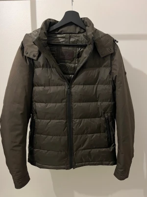 Olivgrön dunjacka från Massimo Dutti - Snygg olivgrön dunjacka från Massimo Dutti med avtagbar huva och dragkedja framtill. Jackan är quiltad, har vind- och vattentätt material samt innerficka med dragkedja. Perfekt för kyliga dagar och har en clean, modern look. Storlek L men passar M!