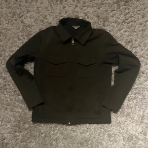 Svart overshirt från NEXT - Svart overshirt från NEXT i storlek XS. Jackan har klassisk krage, två bröstfickor med lock och dragkedja framtill. Långärmad och med stilren, rak passform. Perfekt för dig som gillar minimalistisk och clean look.