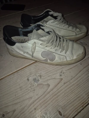 THE GALERIST vita sneakers med svart häl - Snygga vita sneakers från Galliano med svart häl och grå klubb-symbol på sidan. Skorna har rund tå, platt sula och klassisk snörning. Tillverkade i skinn med detaljer i syntet. Perfekta för dig som gillar unik design och streetstyle.