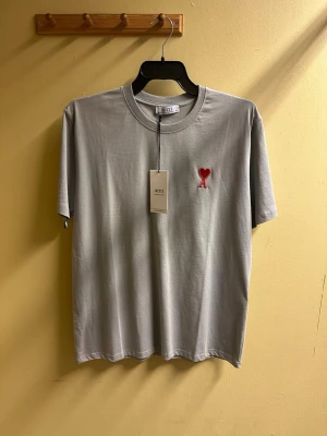 Grå t-shirt från Ami med rött hjärta - Snygg grå t-shirt från Ami Alexandre Mattiussi med klassisk passform. På bröstet finns ett broderat rött hjärta med bokstaven A. T-shirten är i mjuk bomull och har rund hals och korta ärmar. Perfekt för en clean och stilren look.