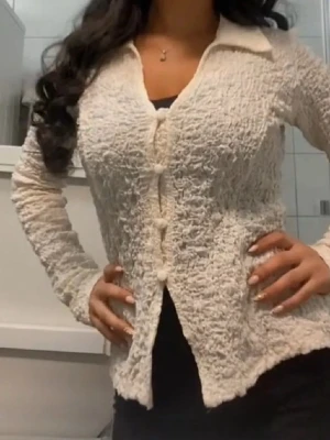 Krämvit kofta från Gina - Säljer en krämvit kofta från Gina  med coolt skrynkligt tyg och stora tygklädda knappar. Koftan har långärmad passform, klassisk krage och är gjord i ett lätt, stickat material som känns mjukt mot huden. Perfekt för dig som gillar unika detaljer och stilren look.