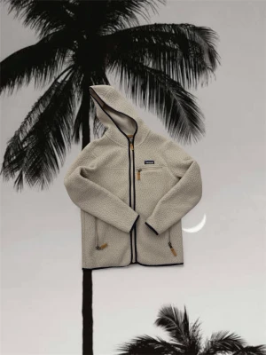 Patagonia fleece - Mysig beige teddyjacka från Patagonia med huva och svarta detaljer. Jackan har tre fickor med dragkedja och snygga bruna zip-pullers. Perfekt för kalla dagar, med långärmad design och mjukt fleece-material som håller dig varm.