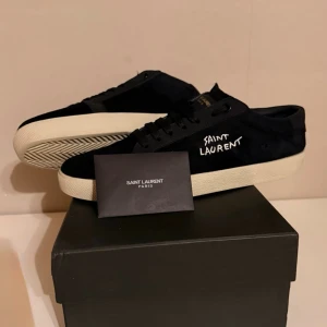 Svarta Saint Laurent sneakers - Svarta sneakers från Saint Laurent med vit handskriven logga på sidan. Skorna har en klassisk låg profil, snörning och beige platt gummisula. Ovandelen är i canvas och detaljer i skinn. Perfekta för en clean och stilren look.