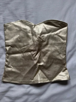 Guldig axelbandslös tubtopp - Snygg guldig tubtopp med glansig finish och hjärtformad överkant. Toppen är axelbandslös och har en dragkedja baktill för enkel på- och avtagning. Perfekt för dig som vill sticka ut med en trendig och stilren look. Aldrig använd så helt ny