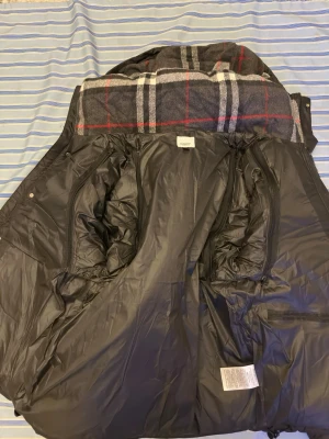 Svart pufferjacka från Burberry - Svart pufferjacka från Burberry med klassisk rutig insida i grått, vitt och rött. Jackan har huva, synliga sömmar och patch med Burberry London England på ärmen. Fodret är quiltat och jackan har dragkedja och knappar framtill.