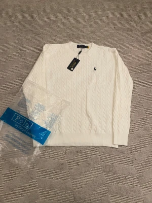Vit kabelstickad tröja Polo Ralph Lauren - Vit kabelstickad tröja från Polo Ralph Lauren med klassisk rund hals och broderad logga på bröstet. Tröjan har långärmade ribbade muddar och är gjord i mjuk bomull. Perfekt för dig som gillar clean och stilren look.41 cm vid axlarna och 62 i längd 