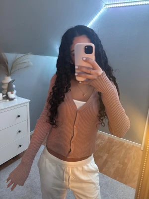 Ribbad beige kofta från H&M - Superfin ribbad kofta från H&M i en dammrosa, lite varm ton. Croppad modell med v-ringning och knappar framtill. Långa ärmar och stretchigt, mjukt material som sitter snyggt på kroppen. Perfekt att styla med linne eller t-shirt under.