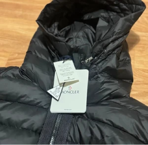 Moncler cardigan - Svart pufferjacka från Moncler med huva och ribbade svarta ärmar. Jackan har Moncler-logga på ärmen och dragkedja framtill. Quiltad design och stilren look, perfekt för dig som gillar exklusiva märken och vill hålla dig varm med stil.