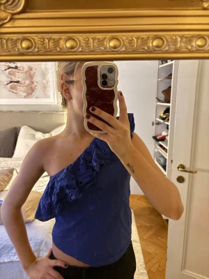 Blå oneshoulder topp med volang - Säljer en blå oneshoulder topp med snygg volangdetalj över axeln. Toppen är ärmlös på ena sidan och har broderade hålmönster i volangen. Perfekt för dig som vill sticka ut med en cool och unik look.