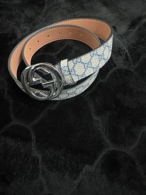 G bälte  - Snyggt g bälte i blått på beige botten. Bältet har en stor, rund silverspänne. Tillverkat i skinn med canvas-yttersida och exklusiv känsla. Perfekt för att lyfta vilken outfit som helst.