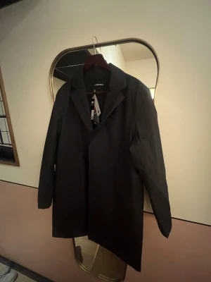 Svart rock från J.Lindeberg, S - Svart, stilren rock från J.Lindeberg i modellen Farris 2L Padded Coat. Jackan har klassisk krage, raka linjer och diskret JL-logga på ärmen. Tillverkad i syntetmaterial med lätt vaddering, perfekt för kyliga dagar. Snygg och enkel att matcha med olika outfits. Ny med taggar