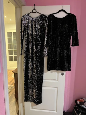 Svart glittrig paljettklänning - Säljer två svarta  glittriga  paljettklänning med rund hals och trekvartsärm. Klänningar  är täckta  av svarta paljetter som ger en riktigt snygg och festlig look. Passformen är normal och materialet känns som polyester. Perfekt för dig som vill sticka ut med lite extra bling. Den långa passar i storlek S och den korta passar i storlek XS.
