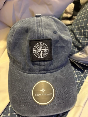 Blå keps från Stone Island - Snygg blå keps från Stone Island med klassisk böjd skärm och broderad logotyp framtill. Kepsen är tillverkad i mjuk bomull och har en stilren look som passar perfekt till streetwear. Justerbar baktill för bästa passform.