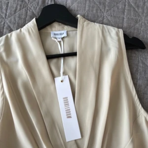 klänning från Bubble Room - Elegant beige klänning från Bubble Room med omlott-design och knytband i midjan. Klänningen är ärmlös och har en v-ringad halslinje, perfekt för en stilren look.