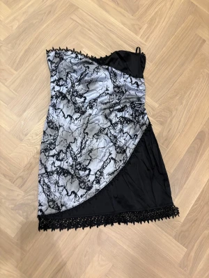 Svart och silver axelbandslös klänning - Axelbandslös kortklänning i svart och silver med marmorerat mönster framtill. Klänningen har spetsdetaljer längs överkant och nederkant samt dragkedja i ryggen. Materialet är glansigt och känns som polyester, vilket ger en cool vibe. Perfekt för dig som gillar unika detaljer och kontraster.