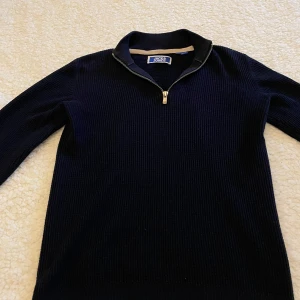 Mörkblå stickad half zip från Jack & Jones - Mörkblå stickad tröja från Jack & Jones med ribbad struktur och halv dragkedja framtill. Hög krage och långärmad modell, perfekt för lager-på-lager. Klassisk och stilren look som funkar året runt.