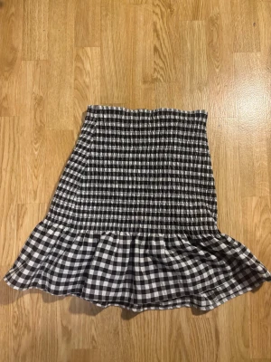 Svartvit rutig volangkjol från New Yorker - Supersöt kort kjol från New Yorker i svartvitt rutigt mönster. Kjolen har smockad midja och en bred volang längst ner som ger en riktigt skön och lekfull vibe. Perfekt för dig som gillar en trendig och bekväm stil.