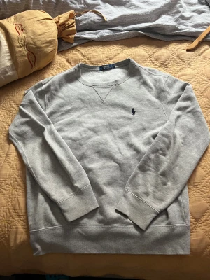 Grå sweatshirt från Polo Ralph Lauren - Säljer en stilren grå sweatshirt från Polo Ralph Lauren i storlek M. Tröjan har långärmade ribbade muddar, rund hals och den klassiska mörkblå Polo-loggan broderad på bröstet. Materialet är mjuk bomull med insida i fleece för extra komfort.