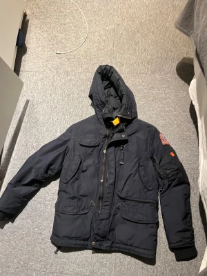 Mörkblå Parajumpers Anchorage jacka - Säljer en svart Parajumpers Anchorage jacka med huva och flera fickor. Den har en defekt vid vänstra armen bild kan fås vid fråga. Jackan har coola patchar på ärmen och insidan, samt en orange detalj vid halsen. Materialet är slitstark polyester och jackan är perfekt för kalla vinterdagar. Grym design och riktigt skön passform. Har 