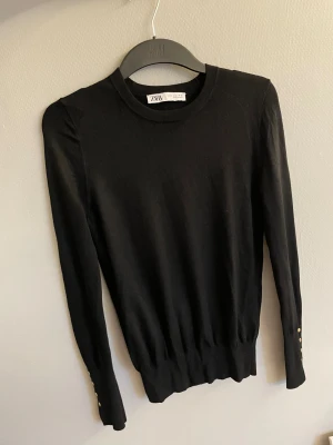 Svart långärmad tröja från Zara - Svart långärmad tröja från Zara med rund halsringning och snygga guldfärgade detaljer vid ärmsluten. Tröjan är ribbad nedtill och har en stilren, enkel design. 