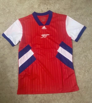 Arsenal röd kortärmad fotbollströja Adidas - Säljer en Arsenal fotbollströja från Adidas i rött med vita och blå detaljer. Tröjan har korta ärmar, blå krage och vita ärmslut. Materialet är lätt och andas, perfekt för match eller träning. Snygga ränder och klubbens kanonlogga på bröstet.