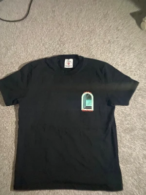 Svart Casa Blanca t-shirt med tryck - Svart t-shirt från Casa Blanca med stort grafiskt tryck av ett fönster i turkos och vitt på ryggen samt mindre tryck på bröstet. Röd text 'Casa Blanca' både fram och bak. Klassisk rund hals och korta ärmar. Skön och mjuk bomullskvalitet.