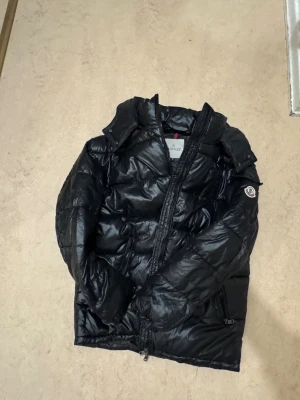 Svart pufferjacka från Moncler M - Svart pufferjacka från Moncler i storlek M. Jackan har en glansig finish, huva med tryckknappar, två snedställda fickor med dragkedja och klassisk Moncler-logga på ärmen. Perfekt för kalla dagar med sin quiltade design och dunfyllning. 