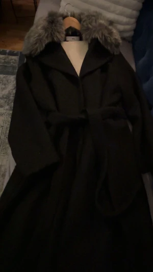 Svart kappa med fuskpäls från Ellos - Snygg svart kappa från Ellos med stor grå fuskpäls på kragen. Modellen är lång med bälte i midjan och har klassisk krage. Perfekt för dig som vill ha en stilren och varm look under vintern.