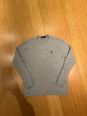 Grå kabelstickad tröja Polo Ralph Lauren - Kabelstickad grå tröja från Polo Ralph Lauren med klassisk rund hals och broderad logga på bröstet. Tröjan är långärmad och har ribbade muddar vid ärmslut och nederkant. Perfekt för lager-på-lager och snygg till jeans.