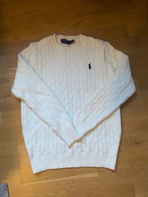 Vit kabelstickad tröja Polo Ralph Lauren - Vit kabelstickad tröja från Polo Ralph Lauren i storlek S. Klassisk rund hals och långärmad modell med broderad marinblå logga på bröstet. Mjuk och skön känsla, perfekt för dig som gillar stilrena och tidlösa plagg.