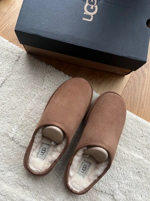 UGG bruna slip-in tofflor med foder - Mysiga slip-in tofflor från UGG i brun mocka med mjukt vitt foder på insidan. Rund tå och platt sula med UGG-logga på sidan. Perfekta för chill hemma och har en stilren, enkel design som passar till allt. HELT NYA! 