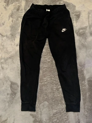 Svarta mjukisbyxor från Nike, storlek S - Svarta mjukisbyxor från Nike med vit broderad logga på vänster ben. Byxorna har elastisk midja med snörning och ribbade muddar vid bensluten. Tillverkade i mjukt bomullsmaterial för extra komfort. Perfekta för chill eller träning.