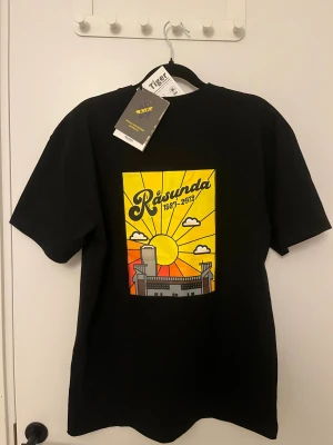 Aik t shirt - Svart t-shirt från AIK med tryck 'Råsunda 1937-2012' i gult på bröstet och en stor färgglad grafik på ryggen med sol, moln och Råsunda stadion. Rund hals och korta ärmar. Officiell merchandise, tillverkad i mjuk bomull. Stor i storlek 