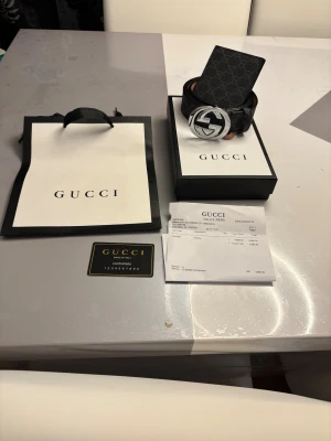 Svart Gucci bälte med GG-spänne - Säljer ett svart Gucci bälte med klassiskt GG-mönster och en stor, rund silverfärgad GG-spänne. Bältet är gjort i äkta skinn och har exklusiv känsla. Kommer med originalförpackning, kvitto och kontrollkort. Perfekt för dig som vill ha en lyxig detalj till outfiten.