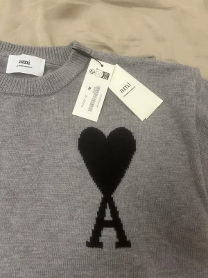 Grå stickad tröja från Ami med hjärta - BLACK FRIDAY! Man kan inte hitta bättre/snyggare material än detta, snygg grå stickad tröja från Ami med svart hjärta och bokstaven A broderat på bröstet. Tröjan har rund hals, ribbade muddar och är långärmad. Perfekt för dig som gillar stilrena detaljer och vill ha något som sticker ut lite extra.