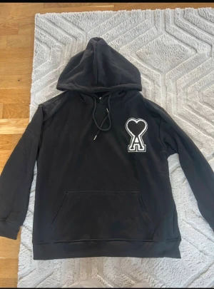 Svart hoodie från Ami Paris med hjärtdetalj - Svart hoodie från Ami Paris i storlek S med stor huva och snörning. På bröstet finns en vit broderad hjärtdetalj med bokstaven A. Tröjan har en klassisk känguruficka och är tillverkad i mjuk bomull. Perfekt för dig som gillar stilrena streetwear-plagg.