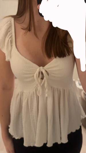 Vit blus med knyt och volang - Supersöt vit blus med knyt framtill och volangdetaljer vid axlarna. Blusen har en lätt rynkad struktur och är luftig med en flowy passform. Perfekt för dig som gillar romantisk och feminin stil. Materialet känns mjukt och lätt mot huden.