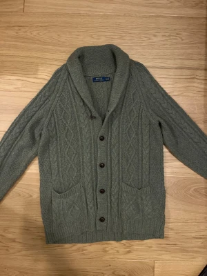 Cardigan från Polo Ralph Lauren XL/L - Olivgrön, grovstickad cardigan i nyskick från Polo Ralph Lauren med snygg kabelstickad design, bred krage och bruna knappar framtill. Två praktiska fickor och långärmad modell. Perfekt för att hålla sig varm och se chill ut under hösten och vintern. Storlek XL passar även L. Priset inte hugget i sten!